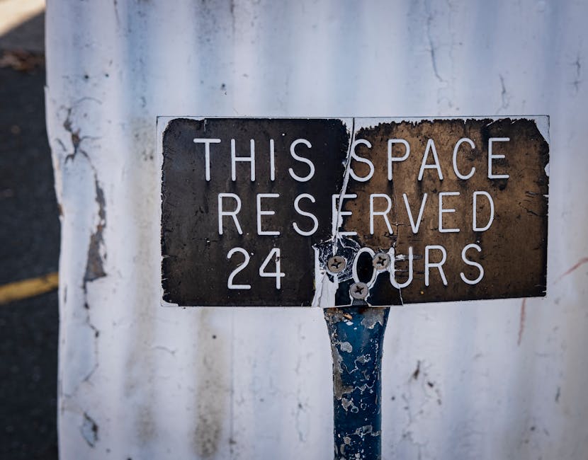A weathered black and white metal sign mounted on a pole, reading 'This space reserved 24 hours,' with the paint chipped and rust visible, attached to a background of a weathered white wall with peeling paint. The sign appears old and slightly damaged, with visible screws and torn paper remnants. This image is relevant to house removals and parking permit regulations, and is displayed on the webpage about avoiding parking fines during Eastcote moves, provided by Man with Van Eastcote, a professional removals company specializing in packing, furniture transport, and home relocation services.