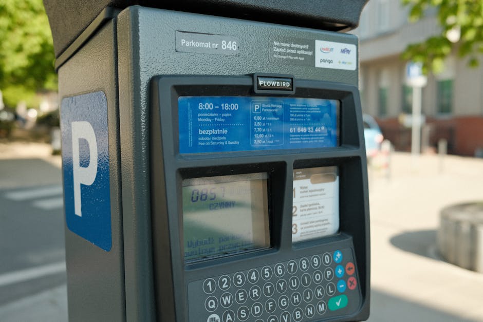 Close-up view of a digital parking meter situated on a paved street in Eastcote, displaying parking time and fee information with a large blue 'P' sign attached. The meter is metallic grey with a touchscreen interface, showing instructions in Polish and timings from 8:00 to 18:00 on weekdays, with free parking on weekends. The surrounding environment includes blurred images of residential buildings with windows and a tree-lined sidewalk, indicating a suburban residential area. The photograph captures the meters during daylight, with natural light illuminating the scene. This setting relates to house removals and moving services by emphasizing the importance of parking management and permits during a home relocation, as managed by Man with Van Eastcote, ensuring movement logistics are efficient and compliant with local parking regulations.