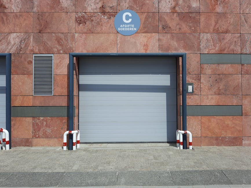 The image shows a closed grey roller shutter door set into a modern exterior wall of a building with reddish-brown stone tiles. On either side of the door, there are short black and white safety bollards positioned on a paved driveway area, designed to protect the entrance from vehicle impacts. Above the door, a circular blue sign with white text reads 'AFGIFTE GOEDEREN,' indicating a commercial or delivery entry point. To the right of the door, there is a small rectangular white sensor or keypad mounted on the wall, likely part of access control for the loading bay. To the left, a vertical ventilation grille is embedded into the wall. The surrounding environment appears clean and well-maintained, with the scene captured during daylight, highlighting the functional loading area suitable for home relocation or furniture transport, as managed by professional removals services such as Man with Van Eastcote.