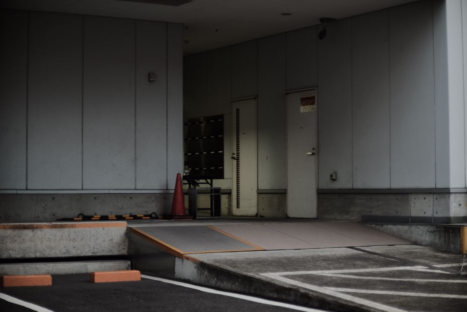 The image shows a driveway loading bay adjacent to a grey industrial or commercial building, with a sloped concrete ramp designed for easy access. The loading bay features a flat, textured metal surface for smooth furniture and box transportation, with orange edging to indicate the ramp's boundary. To the left, there are two orange traffic cones and a black and yellow striped safety barrier on the pavement, which is marked with white lines to designate space for loading and parking. Near the back of the loading area, there is a small metallic table or cart, along with several black individual mailboxes or storage compartments mounted on the building wall. The building's grey paneled exterior includes a small white door, a closed access hatch, and an electrical outlet. The scene is lit with natural light, highlighting the clean, precise environment appropriate for home relocation or furniture transport tasks, which Man with Van Eastcote handles as part of their removals services.