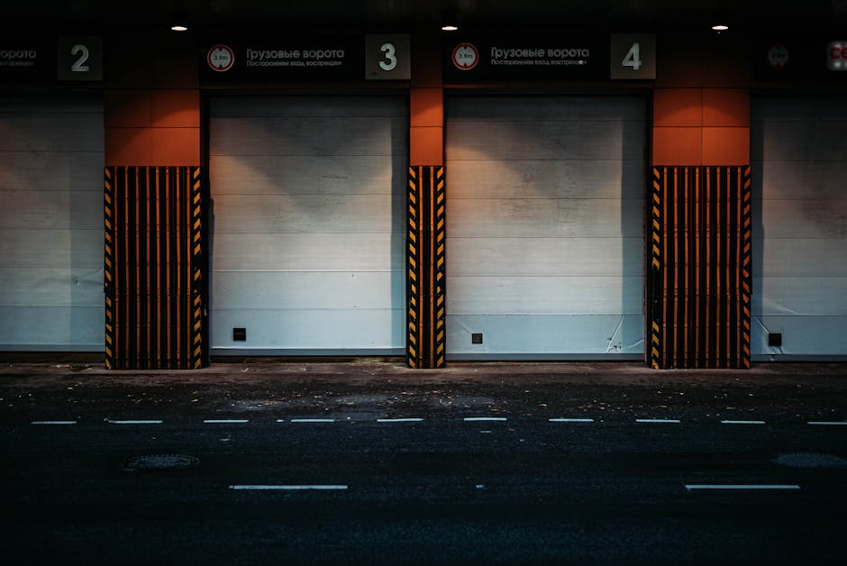 The image shows a closed grey roller shutter door set into a modern exterior wall of a building with reddish-brown stone tiles. On either side of the door, there are short black and white safety bollards positioned on a paved driveway area, designed to protect the entrance from vehicle impacts. Above the door, a circular blue sign with white text reads 'AFGIFTE GOEDEREN,' indicating a commercial or delivery entry point. To the right of the door, there is a small rectangular white sensor or keypad mounted on the wall, likely part of access control for the loading bay. To the left, a vertical ventilation grille is embedded into the wall. The surrounding environment appears clean and well-maintained, with the scene captured during daylight, highlighting the functional loading area suitable for home relocation or furniture transport, as managed by professional removals services such as Man with Van Eastcote.