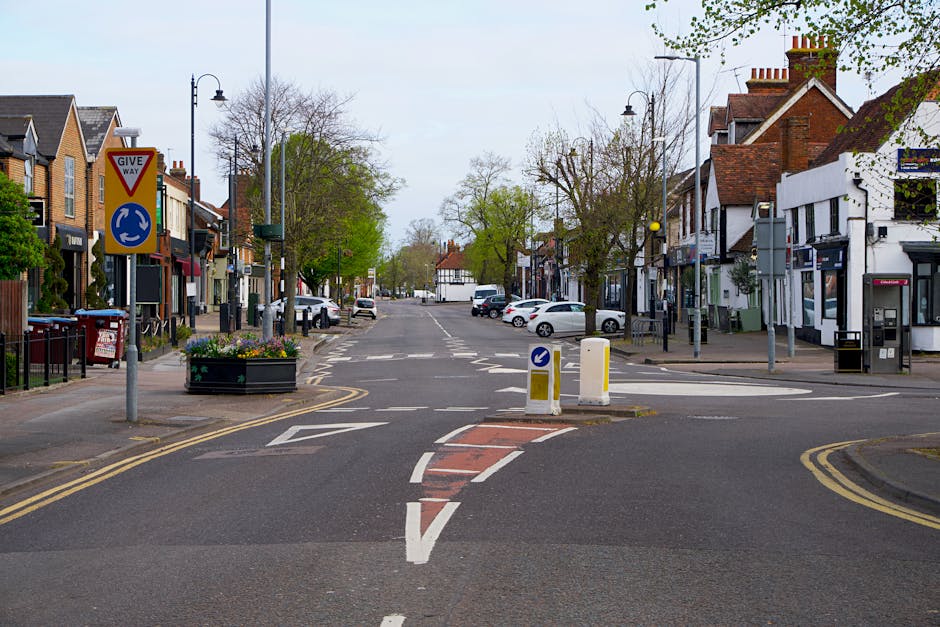 A quiet residential street in Eastcote with a temporary road closure setup, featuring a 'Give Way' sign, a roundabout signage, and road barriers placed in the center of the intersection. On the left s