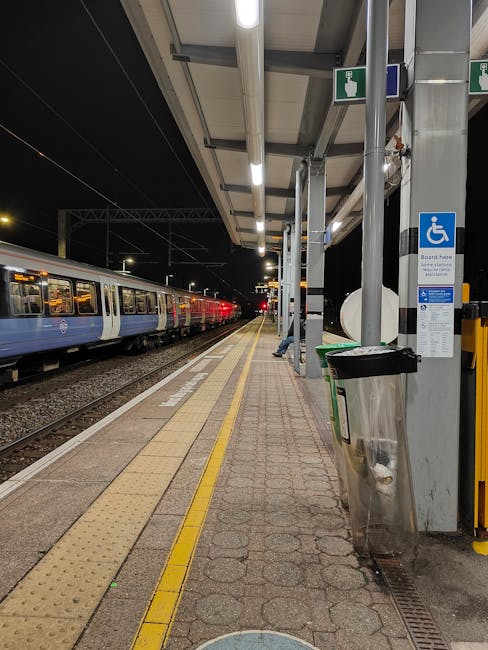 A view of an outdoor train station platform at night with a stationary train on the left side, featuring multiple blue and white carriages illuminated by overhead lighting. The platform surface is paved with textured red brick tiles, with a yellow tactile strip running along the edge for safety. To the right, there is a sheltered waiting area with a roof supported by metal poles. Attached to one pole is a blue and white disability access sign, and beneath it, a black and green trash bin is mounted. Several cardboard boxes and wrapped furniture items are visible on the platform, indicating a home relocation process, as the boxes are stacked near the shelter and a person’s hand is seen lifting or carrying one of them. The background shows darkness with minimal activity, emphasizing the quiet, nighttime setting and the logistics involved in furniture transport and packing for a move. Man with Van Eastcote's removal services are supported by the presence of packed items and the loading environment.