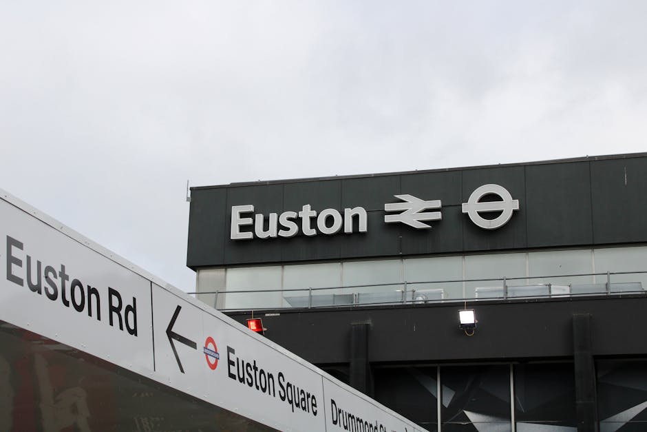 View of Euston station signage against a cloudy sky, featuring the station name 'Euston' alongside the British rail and underground symbols. Below the signage, a white directional sign with black lettering indicates directions to Euston Road, Euston Square, and Drummond Street. The sign is positioned on a pole near the station entrance, suggesting a busy urban environment. This scene relates to house removals and moving services near Euston Station, with the signage providing context for city-based relocation logistics. The photograph highlights the station area, which may be useful for clients planning moving or furniture transport in central London, with the signage serving as a recognizable landmark.