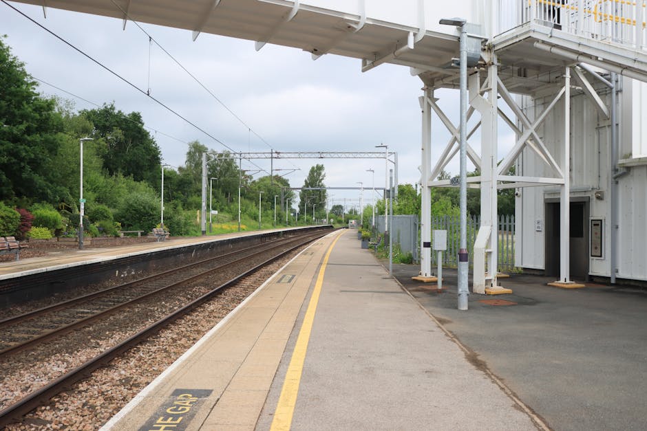 A view of an outdoor train station platform at night with a stationary train on the left side, featuring multiple blue and white carriages illuminated by overhead lighting. The platform surface is paved with textured red brick tiles, with a yellow tactile strip running along the edge for safety. To the right, there is a sheltered waiting area with a roof supported by metal poles. Attached to one pole is a blue and white disability access sign, and beneath it, a black and green trash bin is mounted. Several cardboard boxes and wrapped furniture items are visible on the platform, indicating a home relocation process, as the boxes are stacked near the shelter and a person’s hand is seen lifting or carrying one of them. The background shows darkness with minimal activity, emphasizing the quiet, nighttime setting and the logistics involved in furniture transport and packing for a move. Man with Van Eastcote's removal services are supported by the presence of packed items and the loading environment.