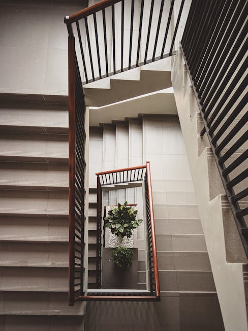 A view looking down a multi-flight staircase in an indoor setting, showing the descending staircases with grey tiled steps, black metal railings on both sides, and wooden handrails. At the bottom of the stairs, there are two potted plants with green foliage placed on the landing. Light from an unseen source illuminates the scene, highlighting the geometric pattern of the staircase. The setting appears to be inside a residential or apartment building in Eastcote, with the staircase arranged in a compact, zigzag layout suitable for house relocations. This arrangement is typical when navigating tight spaces in flats, which may require careful handling during furniture transport and packing and moving processes, as managed by companies like Man with Van Eastcote.