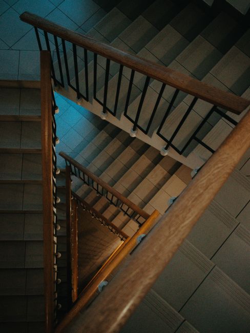 An overhead view of a wooden staircase inside a residential property, with dark metal railings and a polished wooden handrail. The staircase has multiple flights, leading downwards through a narrow corridor with tiled flooring in shades of brown and gray. Several cardboard boxes wrapped in plastic and packing materials are positioned along the staircase, indicating a home relocation process. The lighting appears natural, coming from an unseen source, and the scene is typical of ongoing furniture transport and packing and moving activities. The image is provided by Man with Van Eastcote, a removals service specializing in house relocations and transport logistics.