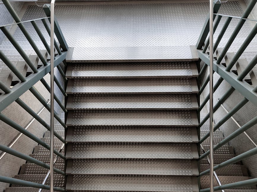 A view looking down a multi-flight staircase in an indoor setting, showing the descending staircases with grey tiled steps, black metal railings on both sides, and wooden handrails. At the bottom of the stairs, there are two potted plants with green foliage placed on the landing. Light from an unseen source illuminates the scene, highlighting the geometric pattern of the staircase. The setting appears to be inside a residential or apartment building in Eastcote, with the staircase arranged in a compact, zigzag layout suitable for house relocations. This arrangement is typical when navigating tight spaces in flats, which may require careful handling during furniture transport and packing and moving processes, as managed by companies like Man with Van Eastcote.