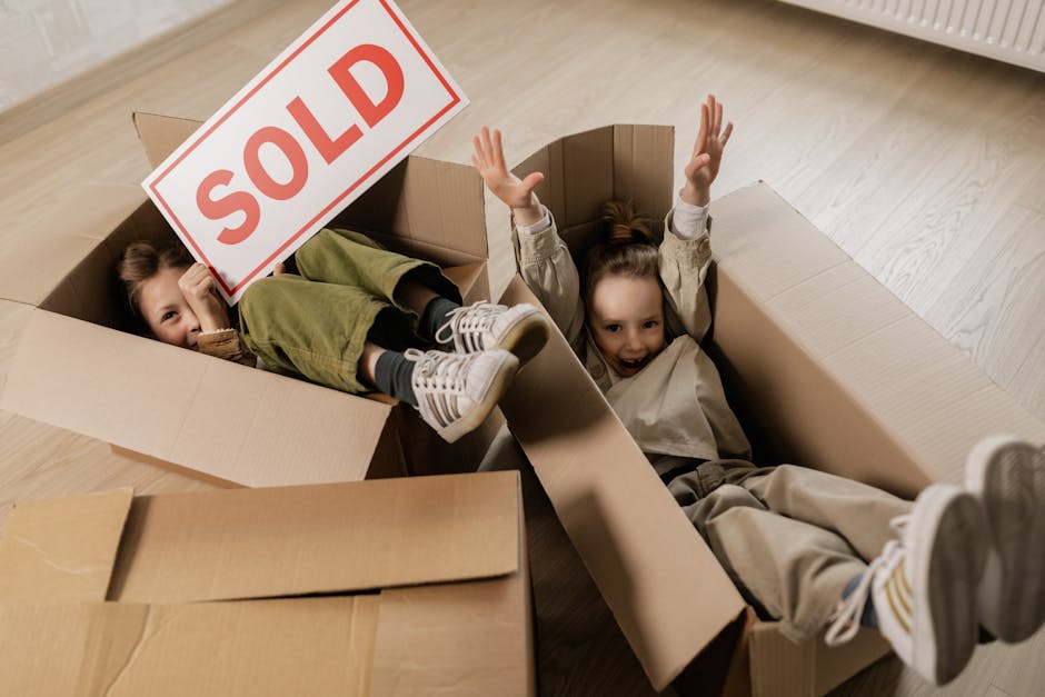 Two young children are lying inside a large open cardboard box, surrounded by other packing boxes on a light wooden floor. One child is partially visible, with their feet sticking out of the box, wearing striped sneakers and green pants, and holding a sign that reads 'SOLD' in red letters. The other child, fully visible, is smiling and has both arms raised above her head, with her legs extended in the box. The scene appears to be indoors, near a white radiator along the wall. This image depicts a playful moment during a home relocation or packing process, which may be managed by professional movers such as Man with Van Eastcote, a company specialising in removals and furniture transport services.
