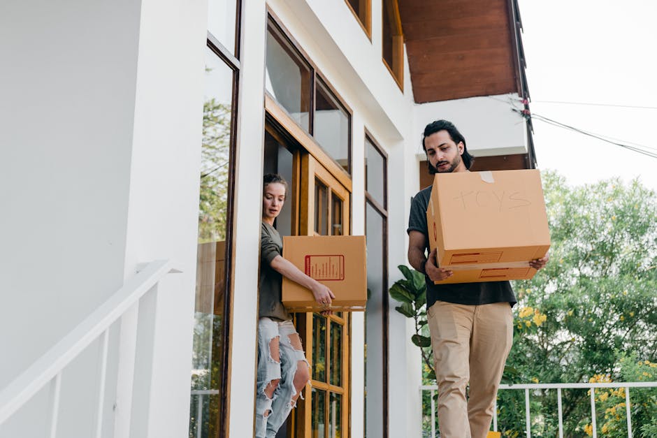 A young man and woman are engaged in a home relocation process outside a residential building, with both carrying cardboard boxes filled with household items through a doorway. The woman, dressed in a casual grey hoodie and ripped jeans, is emerging from the house and holding a box labeled with a red barcode. The man, wearing a dark t-shirt and beige trousers, is standing on a paved outdoor area and holding a larger box marked 'toys.' They are positioned on the steps near the entrance, with interior furnishings and packaging materials likely inside. The background shows greenery and a partly cloudy sky, indicating daytime. The scene reflects the packing and loading process involved in furniture transport and house moving, demonstrating careful handling of household items during a relocation. Man with Van Eastcote offers professional removals, including loading, packing, and transporting services, as depicted in this typical moving scenario.