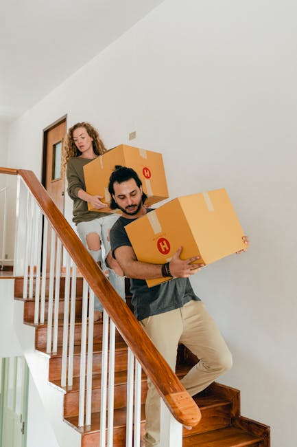 Two movers from Man with Van Eastcote are carrying cardboard boxes down a wooden staircase inside a house. The woman, positioned behind, is holding an open box and wears a beige jacket and ripped jeans. The man, in front, is holding a sealed cardboard box with both hands, wearing a grey T-shirt and beige pants. The staircase features wooden handrails and steps, with a white wall on the side. The scene depicts the home relocation process, including packing and furniture transport, as part of a staircase moving checklist for house removals in Eastcote.