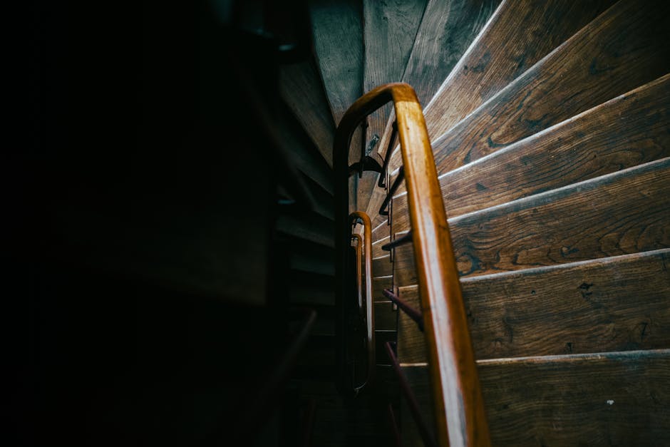 A view from above showing a wooden staircase inside a property, with dark side walls and a polished wooden handrail. The staircase's treads are made of dark, varnished wood, extending downward, with natural wood grain visible. The lighting is dim, highlighting the smooth finish of the wood and the slight reflections on the surfaces. The staircase appears to be in a narrow space, possibly during a home relocation process. Metal support brackets are attached to the under-side of the stairs, and the surrounding walls are painted dark, contrasting with the bright wooden features. This scene depicts part of the moving or packing process, with no visible boxes or moving equipment currently in the image. Man with Van Eastcote's professional removal services focus on safe, efficient furniture transport and staircase disassembly, as shown in the careful handling of interior structures during house removals, especially on staircases like this one.
