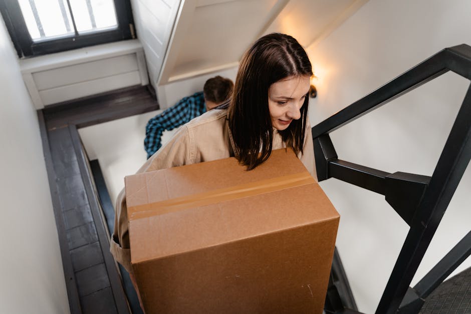 Two movers from Man with Van Eastcote are carrying cardboard boxes down a wooden staircase inside a house. The woman, positioned behind, is holding an open box and wears a beige jacket and ripped jeans. The man, in front, is holding a sealed cardboard box with both hands, wearing a grey T-shirt and beige pants. The staircase features wooden handrails and steps, with a white wall on the side. The scene depicts the home relocation process, including packing and furniture transport, as part of a staircase moving checklist for house removals in Eastcote.